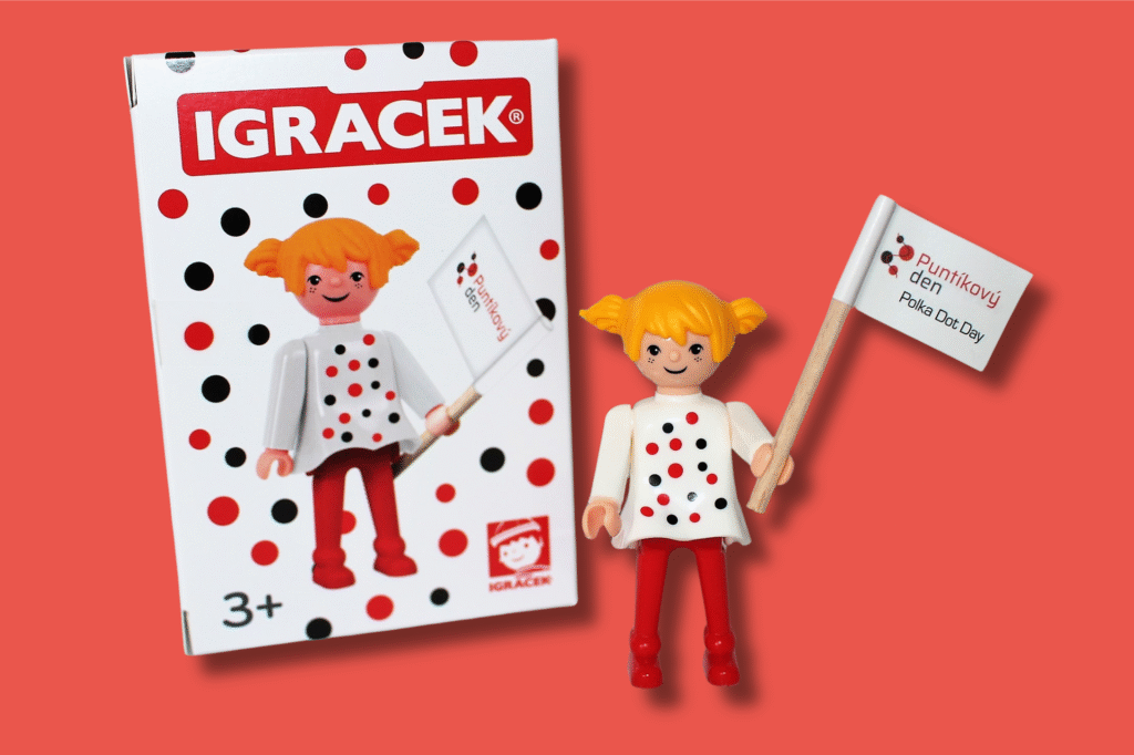 Igráček s puntíky: panenka igráčka v puntíkovaných šatičkách drží v ruce vlaječku s logem Puntíkového dne. Za ní stojí krabička s jejím obrázkem a nápisem "Igráček"